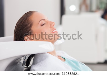 Beautiful woman is having her hair washed in Hair salon. She is sitting and leaning her head on the sink. Her eyes are closed with pleasure. She is smiling