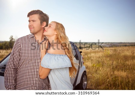 Handsome young man and beautiful woman are enjoying their trip and smiling. They are standing near a car and embracing. The woman closed her eyes with relaxation. Copy space in right side