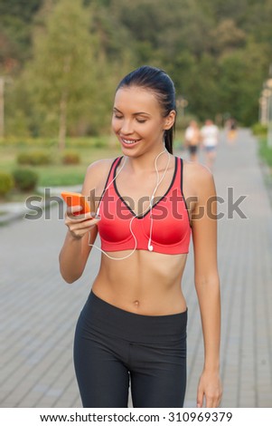 Cheerful young sport woman is running in park. She is holding a mobile phone and looking at it with joy. The girl is messaging to someone and smiling. She is carrying earphones on her neck
