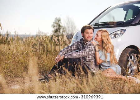 Attractive man and woman are sitting on grass near a car and relaxing. They are looking at each other with love and smiling. The woman is embracing her boyfriend gently. Copy space in left side