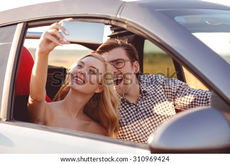 Cheerful young man and woman are sitting in car and making selfie. They are looking at the mobile phone and laughing. The man is driving a vehicle
