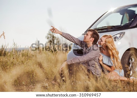 Pretty young loving couple is sitting on grass near a car. The man is pointing his finger sideways with seriousness. The woman is embracing him and smiling. Copy space in left side