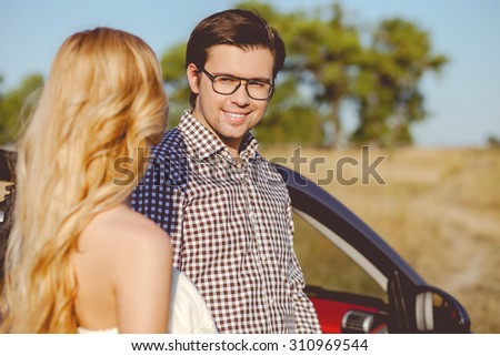 Attractive man and woman are enjoying the nature. They are standing near a car and smiling. The lovers are looking at each other with love