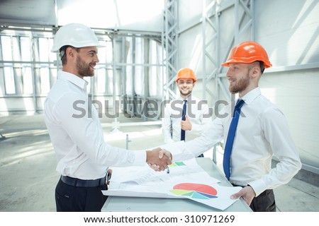 Attractive architects have built a consensus concerning the plan of building. They are shaking hands and smiling. The foreman is standing near them and giving his thumb up with joy