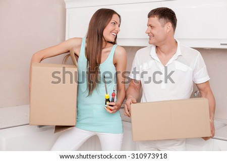 Pretty husband and wife are packing their staff for moving in another house. They are carrying cardboard boxes and smiling. The woman is holding screwdriver and pliers. She is looking at man with joy