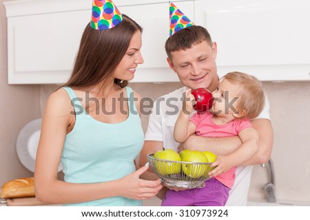 Cheerful family is making fun in kitchen. A man is holding his daughter gently. The child is eating apple with enjoyment. The husband and wife are looking at her and smiling