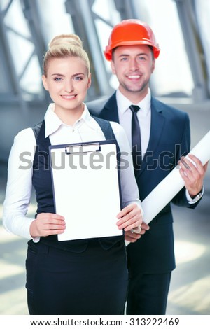 Successful architects are ready to sign a contract with their customer. They are standing and smiling. The woman is showing a folder of documents. The man is holding a blueprint