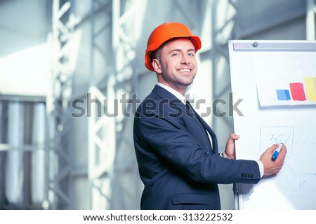 Attractive architect is drawing sketches of construction. He is standing near a board of a blueprint and smiling. He is looking at the camera with joy. Copy space in left side