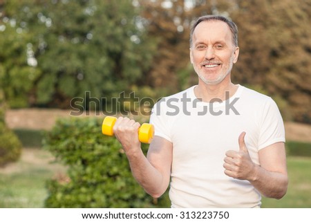 Cute mature sportsman is holding a yellow dumbbell. He is training and smiling. He is standing and giving thumb up. Copy space in left side