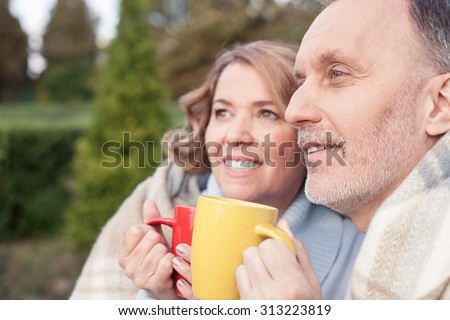 Beautiful old married couple is sitting and relaxing in park. They are holding cups and drinking tea with pleasure. The man and woman are wearing warm quilt and smiling