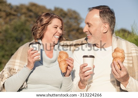 Pretty senior husband and wife are eating croissants in park. They are holding cups and drinking coffee. The family is looking at each other and smiling