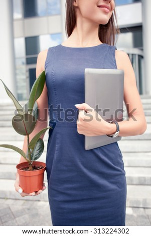 Close up of female body standing on steps neat the office. The woman is holding  a flowerpot and laptop in both her arms. She is smiling