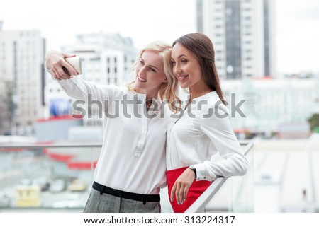 Beautiful female colleagues are standing on balcony of their office. They are making selfie and smiling. The women are embracing and looking at the mobile phone with joy