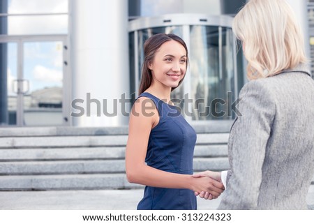 Attractive two businesswomen are shaking hands with joy. They are standing near the office and smiling. The women have built a consensus. Copy space in left side
