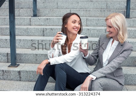Attractive female colleagues are sitting on steps neat their office. They are talking and smiling. The women are drinking coffee with pleasure