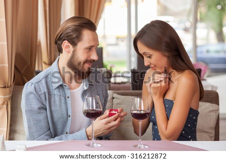 Attractive man is proposing marriage to his girlfriend in restaurant. He is holding a small box of ring and looking at her with hope. They are sitting and smiling. There are two wineglasses on table