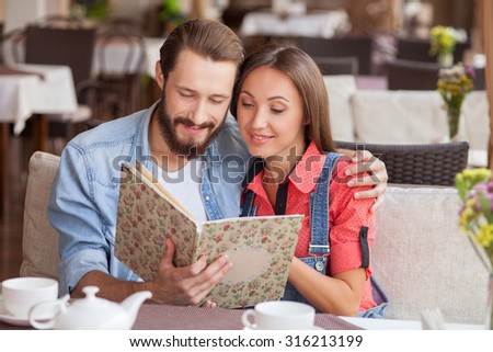 Cheerful man and woman are sitting in cafeteria. They are holding a menu and choosing food. The pair is embracing and smiling