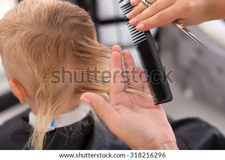 Close up of hands of beautician. The woman is holding scissors and comb. She is cutting hair of child. The boy is sitting on chair. Focus on back of his head