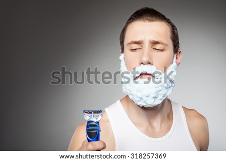 Attractive hipster guy is holding a razor and looking at it with doubt. He has shaving foam all over his beard. Isolated on grey background and copy space in left side