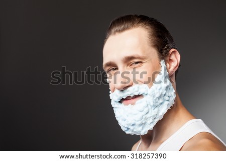 Cheerful man is shaving his beard with joy. He is applying shaving foam on his face. The man is standing and smiling. Isolated on grey background and copy space in left side