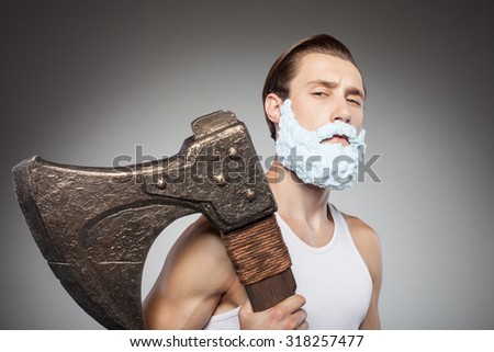 Be afraid of me. Handsome bearded hipster guy is holding big hatchet. He is standing with foam on his beard and looking at the camera with threats. Isolated on grey background