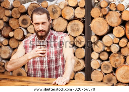 Handsome young man with beard is drinking beer in pub. He is sitting at table and holding glass of lager. The man is looking at camera with confidence. Copy space in right side