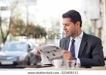 Attractive man in suit is sitting at table in cafe outdoors. He is reading newspaper with interest and smiling. The worker is drinking tea. Copy space in left side
