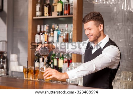Cheerful young bartender is pouring mixed drink from shaker into glasses. He is standing and holding glass. The man is looking at beverage and smiling