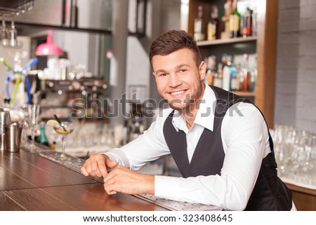 Attractive bartender is standing and leaning on counter is pub. He is looking forward with joy. The man is uniform is smiling