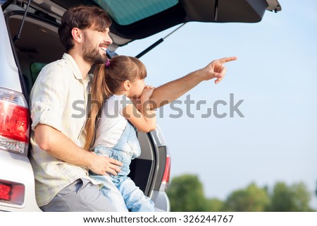 Handsome young father and his small daughter are sitting on open car boot in forest. The parent is pointing finger sideways and smiling. Girl is looking there with interest. Copy space in right side