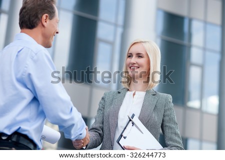 Professional architect and businesswoman are making handshake. They are standing near building and looking at each other with joy. The woman is holding documents. They are smiling