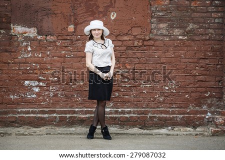 Photo of a woman in a white hat, blouse and black skirt, standing against the backdrop of an old vintage brown brick wall. Space for text.