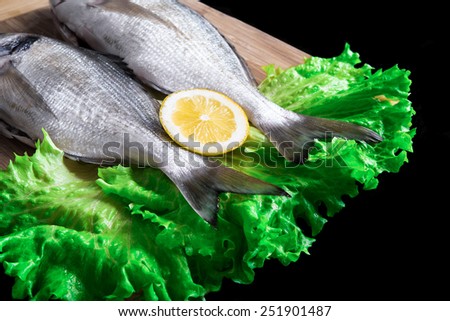 Two fresh dorados on wooden board with green lettuce and slice of lemon isolated on black