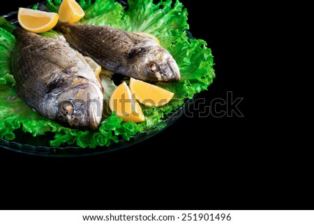 Baked dorados on glass plate with green lettuce and lemons isolated on black