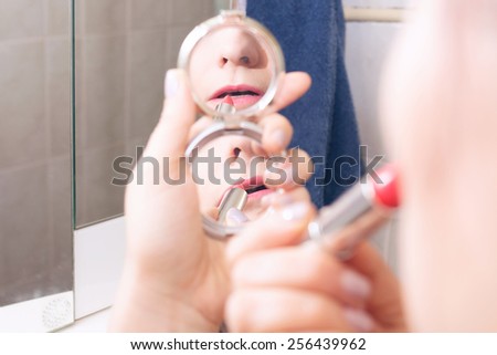 Woman applying lipstick in a bathroom looking in small pocket mirror, focus is on the reflection in the mirror
