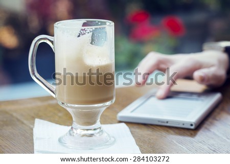 Latte on a table in a cafe on the background of a hand on the tablet. It works in a cafe