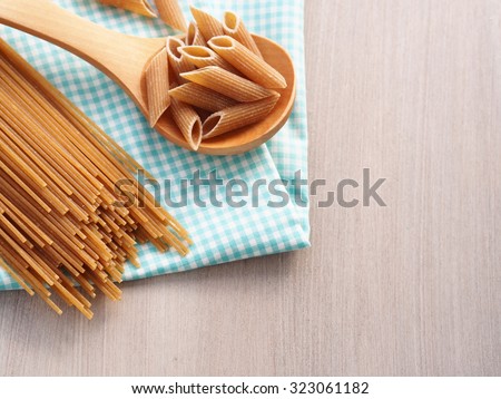 Whole wheat pasta - spaghetti and short pasta penne in wooden spoon on checkered table cloth on wooden table, copy space