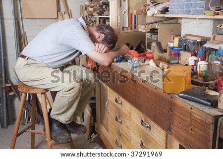 Older man with head on arms in his workshop surrounded by tools and equipment