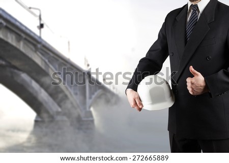 Close up of engineer hand holding white safety helmet for workers security, giving thumbs up success sign, standing in front of blurred urban background.