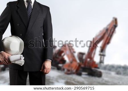 Close up of engineer hand holding white safety helmet for workers security standing in front of  blurred construction site with cranes in background