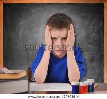 A young boy is sitting at a school desk looking tired and stressed from the lesson. There is a chalkboard in the background for a learning or education concept.