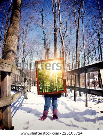 A child is standing on a winter trail in the woods holding a wooden frame with a picture of the summer with sunshine for a season concept.