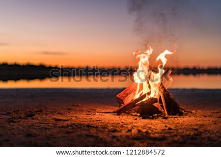 Small campfire with gentle flames beside a lake during a glowing sunset. Western Australia, Australia.