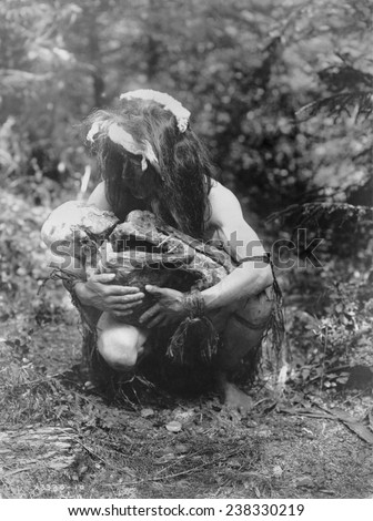 Kwakiutl man, crouched, cradling mummy bound in fetal position, title: Preparing to eat the mummy\', photograph by Edward S. Curtis, circa 1911.