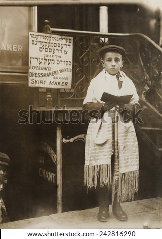 Jewish boy wearing a prayer shawl and boy reading from a Hebrew Bible on Yom Kippur, on a New York City street in a Jewish neighborhood. Ca.1911.