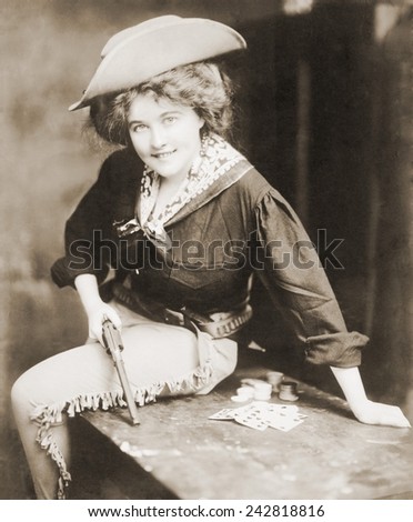 Turn of the century Cowgirl holding revolver, seated on a table, with playing cards and chips. Ca. 1900.