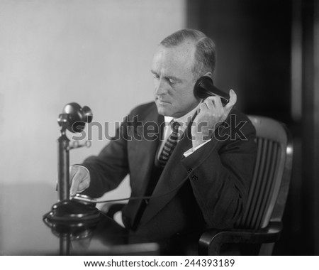 Businessman dialing an early 20th century desk phone with separate ear receiver and mouth transmitter. Ca. 1920.
