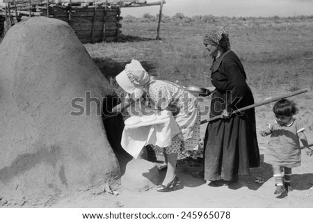 Two Spanish-American women placing loaves of bread in outdoor earthen oven for baking, Taos County, New Mexico