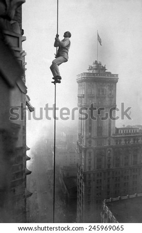 A man, identified as \'the human squirrel\', climbing at a dizzy height above Times Square to promote War Relief Fund contributions. WWI. Ca. 1918.