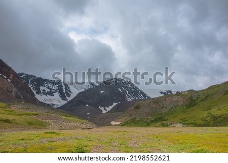 Dramatic alpine scenery with green mountain valley and snow mountains under cloudy sky in changeable weather. Atmospheric overcast landscape with snow mountain range and green valley in sunlight. Foto d'archivio © 